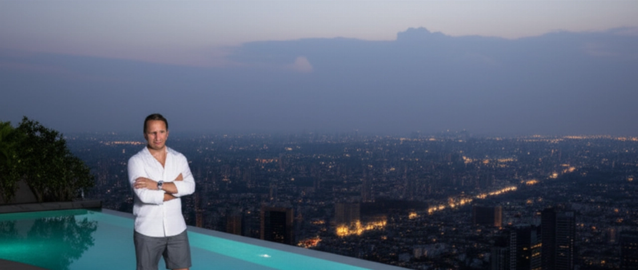 Un uomo caucasico con un Patek Philippe Aquanaut al polso, in piedi accanto a una piscina a sfioro con vista su una città al tramonto.